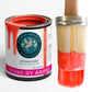 A can of Neon Red Chalk Paint and Clay Paint with a coral undertone, next to a paintbrush with the paint applied to it.