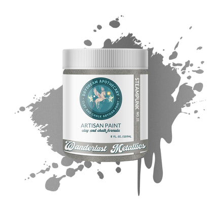 A jar of Wanderlust Metals Artisan Paint in the Steam Punk shade, displayed against a white background with a gray paint splatter graphic. The jar features a white label with a teal circular logo and text.